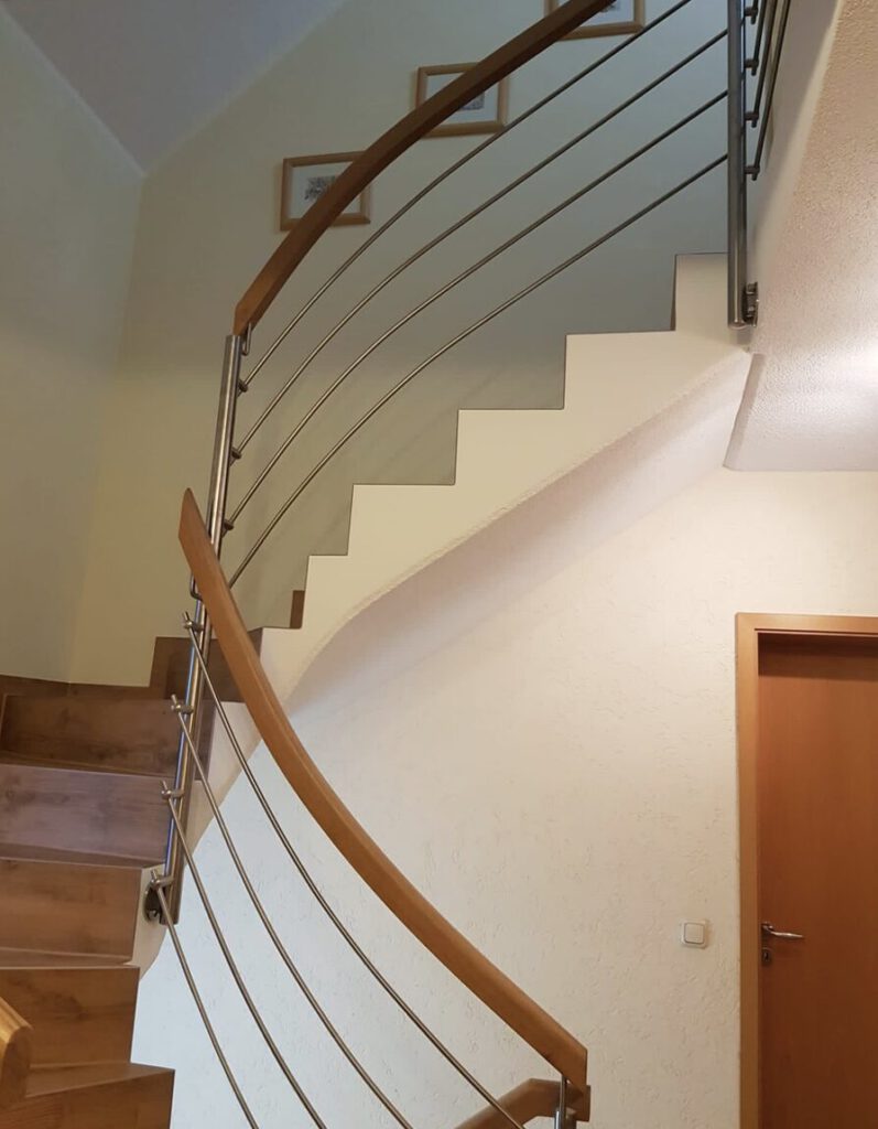 Moderne Innentreppe mit Holzstufen und einem schlanken, geschwungenen Handlauf aus Metall und Holz. Die Treppe führt neben einer hellen Wand hinauf, an der gerahmte Bilder und eine Holztür zu sehen sind.