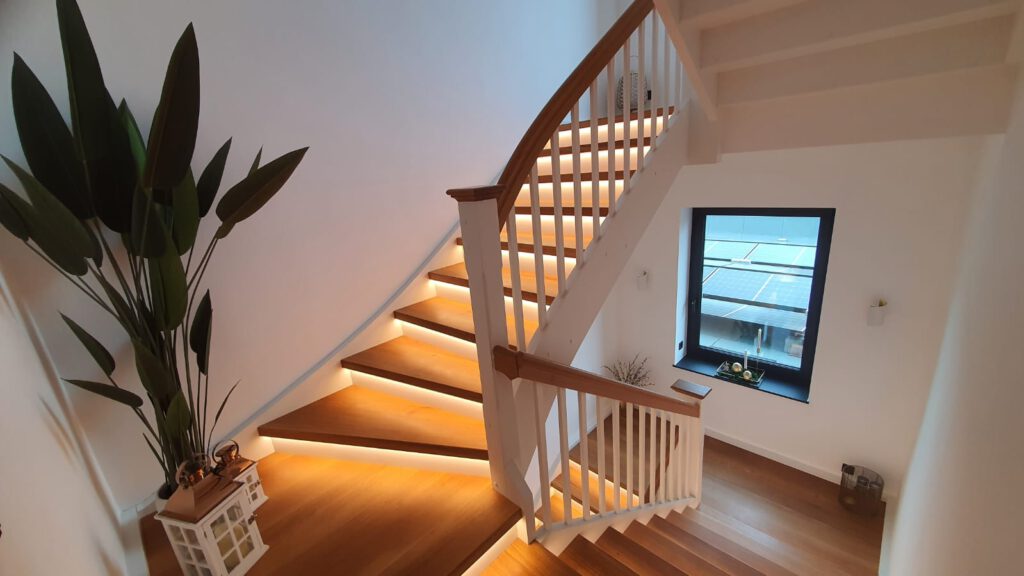 Eine moderne Holztreppe mit warmer, eingebauter Stufenbeleuchtung schlängelt sich neben einer großen Zimmerpflanze und einer dekorativen Laterne nach oben. An der Wand befindet sich ein Fenster mit Blick nach draußen, das den Raum mit natürlichem Licht füllt.