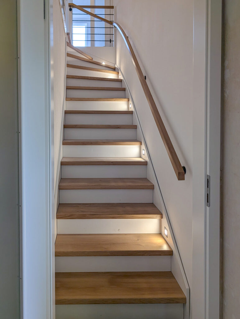 Eine moderne Holztreppe mit eingebauten LED-Leuchten an den Seiten, weißen Wänden und einem hölzernen Handlauf führt zu einem hell erleuchteten Treppenabsatz mit Fenstern.
