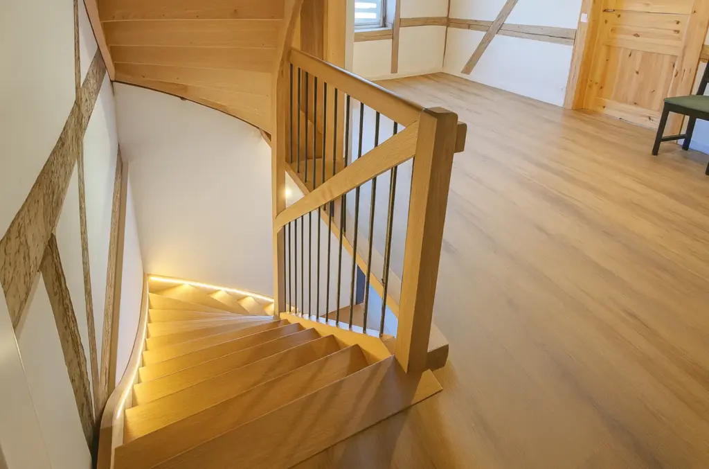 Eine hölzerne Wendeltreppe mit einfachem Geländer führt von einem hellen Raum mit Holzfußböden und -wänden mit sichtbaren Balken und einer Tür auf der rechten Seite hinunter.