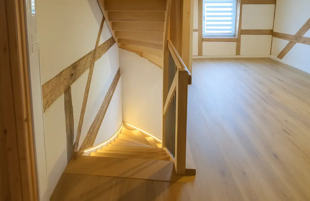 Ein modernes, helles Interieur mit einer Holztreppe mit eingebauter Beleuchtung führt in einen offenen Raum mit Holzböden, sichtbaren Holzbalken, weißen Wänden und einem Fenster mit Jalousien.