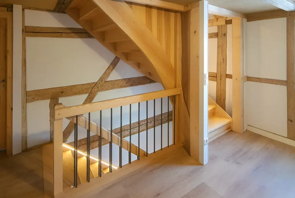 Holztreppe mit offenen Setzstufen und vertikalem Metallgeländer in einem hellen Raum mit sichtbaren Holzbalken, hellen Holzböden und weißen Wänden. Das Licht fällt von unten auf die Treppe und erhellt den Raum.