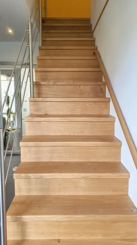 Eine Holztreppe mit breiten Stufen und Metallgeländer auf der linken Seite, die in einen Flur mit gelben Wänden führt. Die Treppe befindet sich in einem Innenraum, in den von links natürliches Licht fällt.