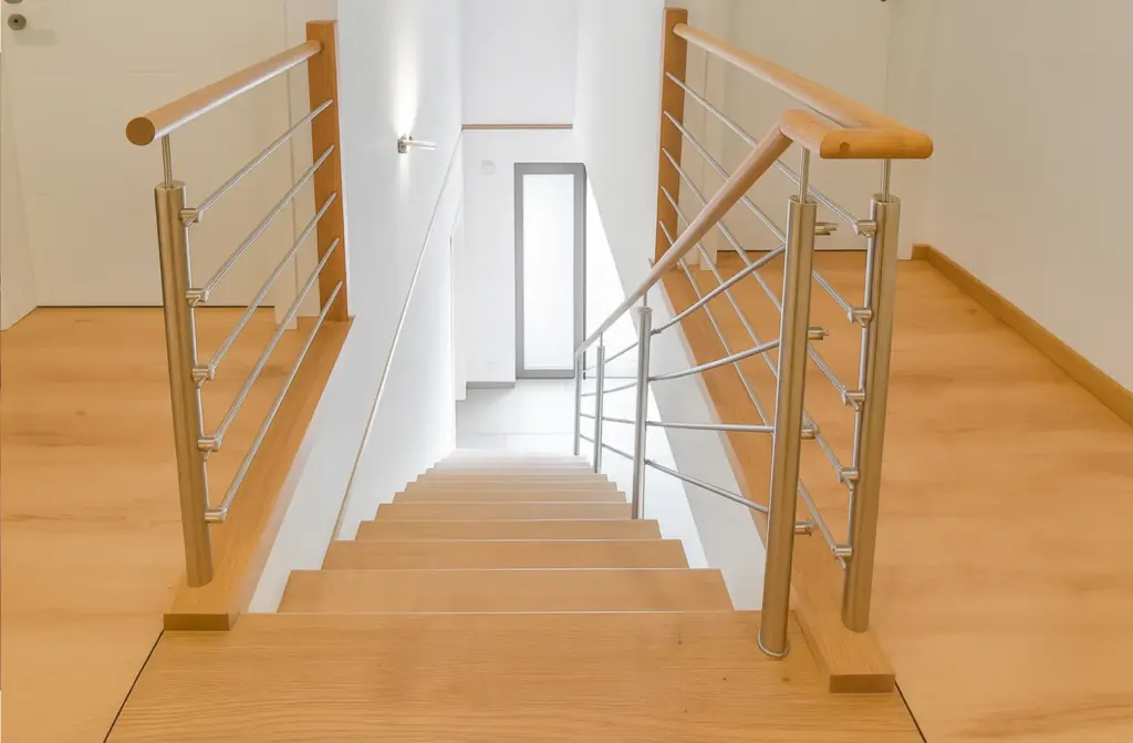 Eine moderne Innentreppe aus Holz mit Metallgeländer führt zu einer hellen unteren Etage mit weißen Wänden und einer unten sichtbaren Tür.