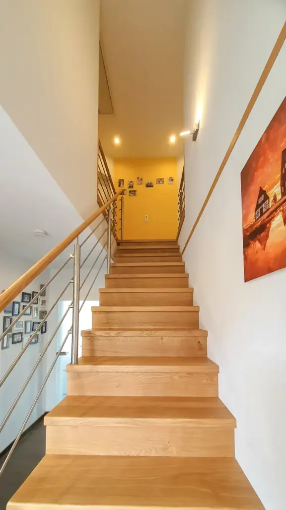 Eine Holztreppe mit Metallgeländer führt in einen Flur mit gelben Wänden. An der gelben Wand und entlang der Treppe sind gerahmte Fotos angebracht, und auf der rechten Seite hängt ein großes Kunstwerk. Der Raum ist gut beleuchtet.