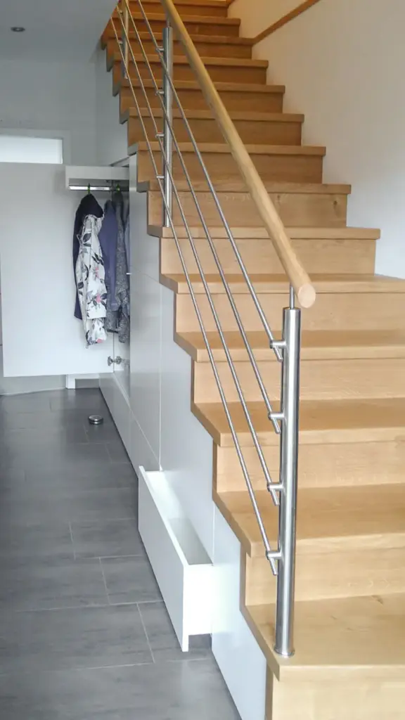 Eine moderne Holztreppe mit Metallgeländer wird neben einem Flur mit grauem Fliesenboden gezeigt. Mäntel hängen an einem Regal unter der Treppe, und ein Futternapf für Haustiere steht an der Wand.