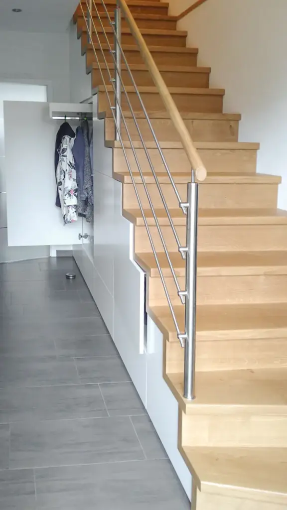 Eine moderne Innentreppe mit Holzstufen und einem Metallgeländer, neben einer weißen Wand. Unterhalb der Treppe befindet sich ein Schrank, in dem Mäntel hängen und dessen Tür offen steht. Der Boden ist grau gefliest.