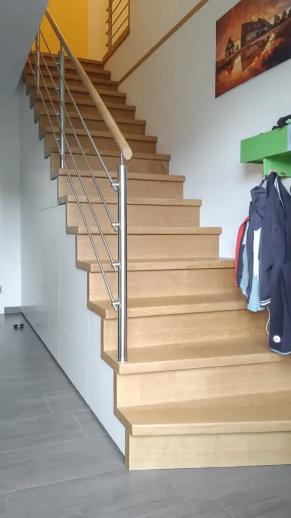 Eine moderne Holztreppe mit einem Handlauf aus Metall, die in ein höheres Stockwerk führt. Rechts hängen Mäntel an Haken, und an der Wand hängt ein Gemälde über einem grünen Regal. Der Boden ist mit grauen Fliesen belegt.