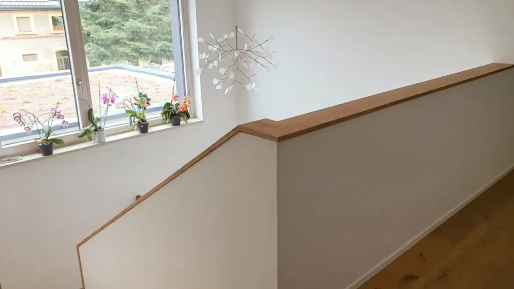 Eine moderne Innentreppe mit einem kurzen Holzgeländer an einer weißen Wand, mehreren Topfpflanzen auf einem Fensterbrett und einem geometrischen Kronleuchter an der Decke.