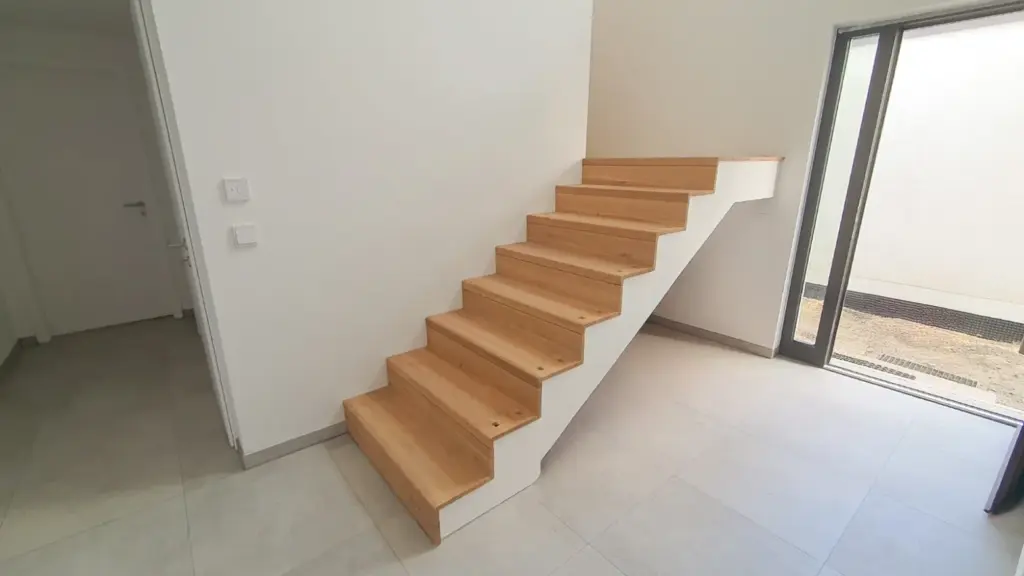Eine einfache Holztreppe mit weißen Wangen in einem modernen, minimalistischen Raum mit hellem Fliesenboden und weißen Wänden. Es gibt eine Tür nach draußen, die natürliches Licht hereinlässt.