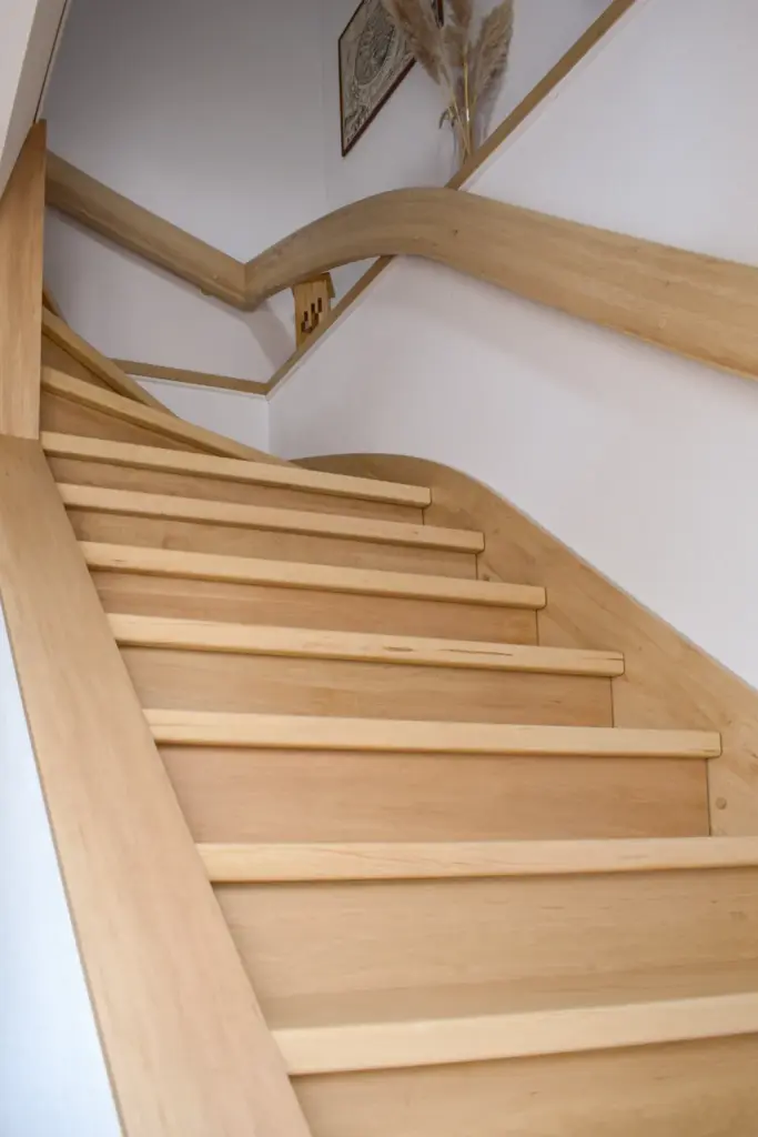 Eine Holztreppe mit glatten, geschwungenen Handläufen führt zwischen weißen Wänden hinauf in ein höheres Stockwerk. Am oberen Ende der Treppe sind ein Dekor aus Pampasgras und gerahmte Kunstwerke zu sehen.