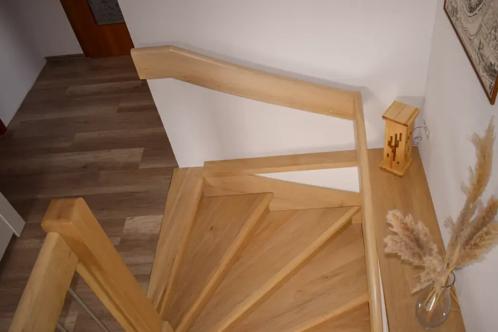 Innentreppe aus Holz mit hellen Stufen und Handläufen, die zu einem Holzboden hinunterführt. Eine Vase mit Pampasgras und eine dekorative Laterne stehen auf einem kleinen Podest neben der Treppe.