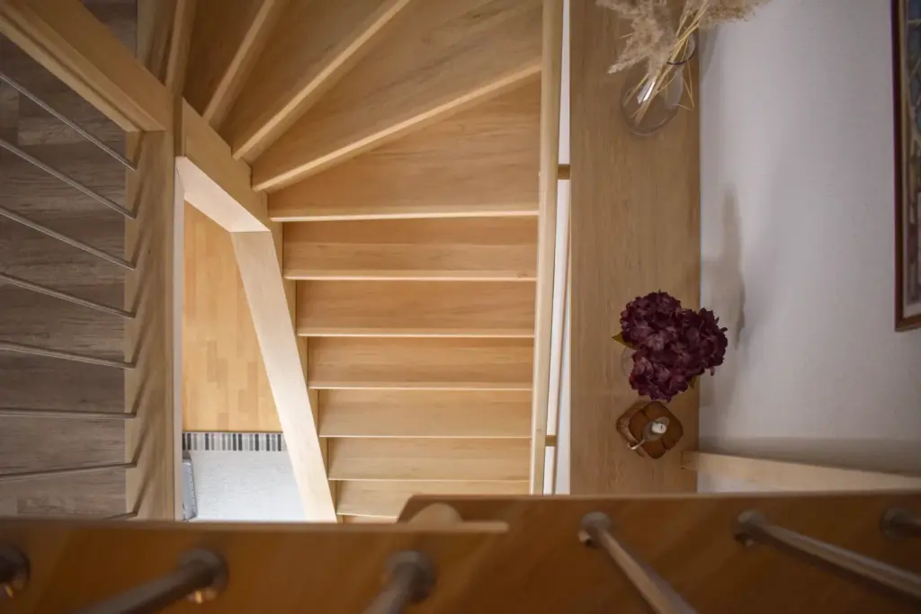 Blick von oben auf eine Holztreppe mit hellen Stufen. Auf einem Podest neben der Treppe ist ein dekorativer Tisch mit einer Vase mit Trockenblumen und einem violetten Blumengesteck zu sehen.
