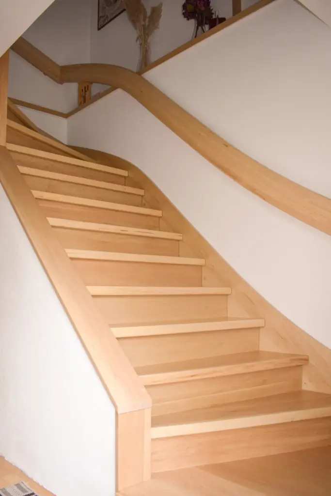 Eine helle Holztreppe mit geschwungenen Handläufen führt entlang einer weißen Wand in einem modernen Innenbereich nach oben. Auf dem oberen Treppenabsatz sind dekorative Pflanzen und gerahmte Kunstwerke zu sehen.