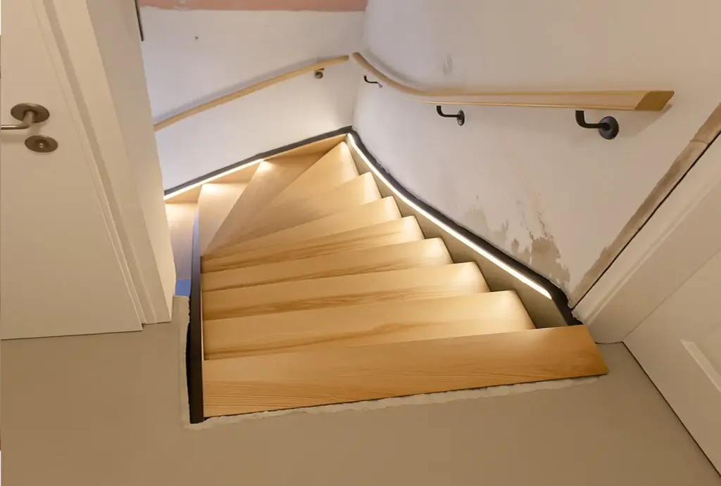 Eine moderne Holztreppe mit beleuchteten Stufen führt steil nach unten, begrenzt von weißen Wänden und einem hölzernen Handlauf auf der rechten Seite; die Wand weist in der Nähe der Treppe einige unfertige Stellen auf.