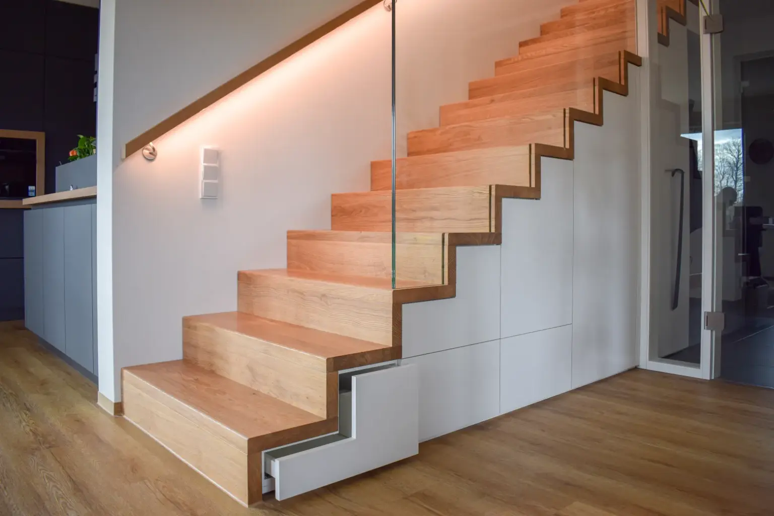 Eine moderne Innentreppe aus Holz mit eingebauter LED-Beleuchtung entlang der Wand, Glasgeländer und weißer Verkleidung darunter, in einem modernen Haus mit Holzfußboden.