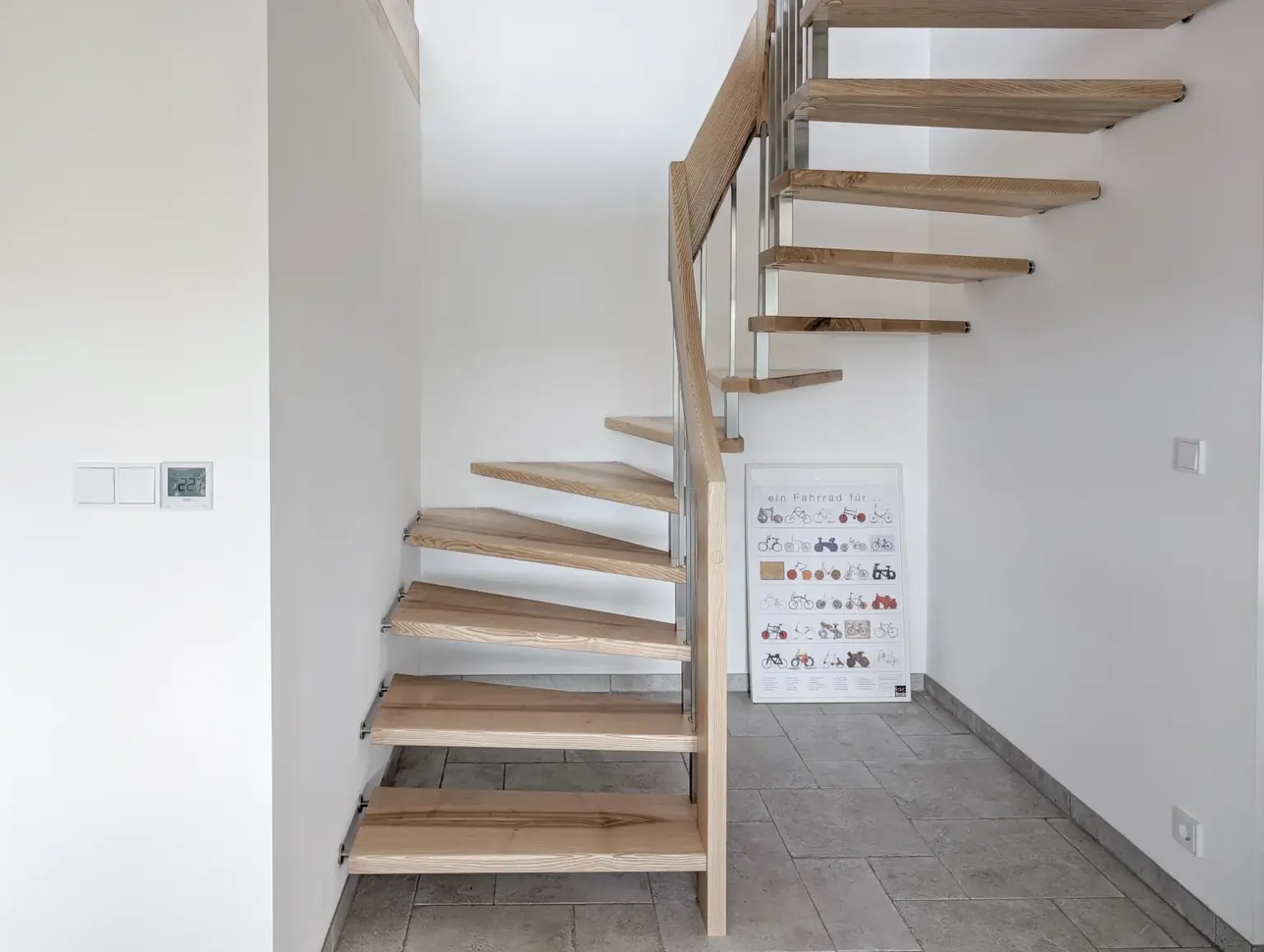 Eine moderne schwebende Holztreppe mit offenen Setzstufen, Metallgeländer und gefliestem Boden in einem hellen, minimalistischen Interieur. An der weißen Wand in der Nähe der Treppe ist eine Tabelle oder ein Poster mit Illustrationen angebracht.