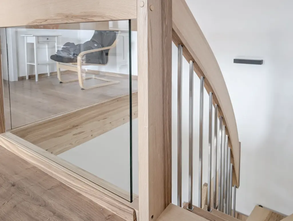 Helle Holztreppe mit vertikalen Metallbalustern und einer Glasscheibe, die in einem modernen, hellen Innenraum nach unten führt. Im Hintergrund befinden sich ein Holzboden, ein weißer Schreibtisch und ein schwarzer Sessel.