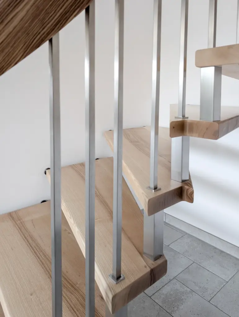 Eine Nahaufnahme einer modernen Treppe mit hellen Holzstufen und vertikalen Metallgeländern vor einer weißen Wand und einem gefliesten Boden.