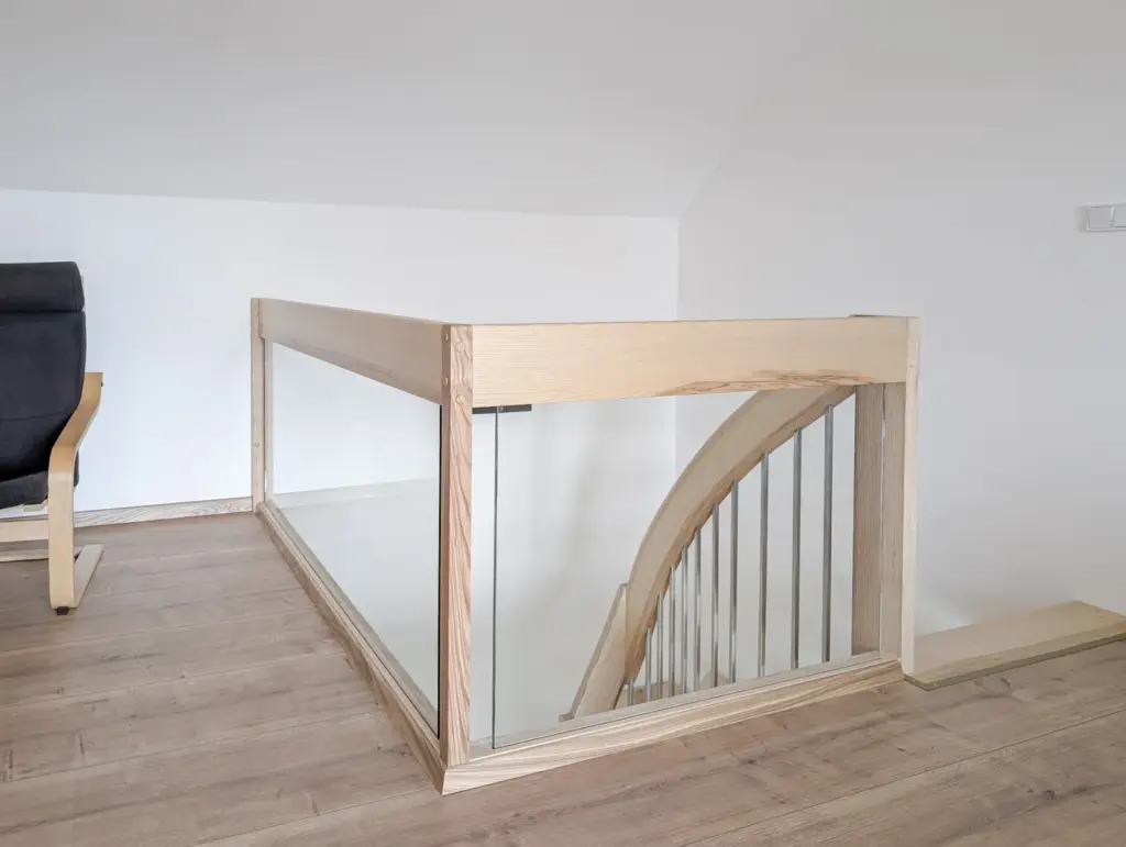 Gezeigt wird eine moderne Innentreppe mit Holzhandläufen und Glaspaneelen. Die Treppe ist nach unten gebogen, und der Boden und die Wände in der Umgebung sind in hellen Farben gehalten, was einen klaren, minimalistischen Look erzeugt. Links ist teilweise ein schwarzer Stuhl zu sehen.