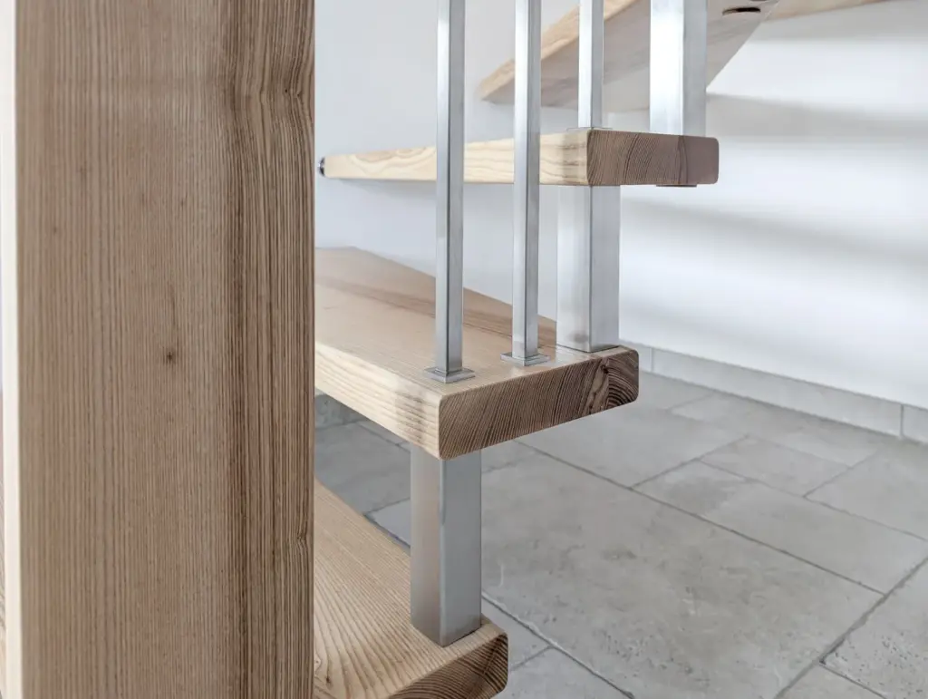 Nahaufnahme einer modernen Holztreppe mit Metallgeländer vor einer weißen Wand und einem hellen Fliesenboden. Das Design wirkt minimalistisch und zeitgemäß.