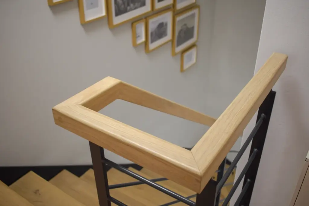 Ein hölzerner Handlauf mit einer quadratischen Ecke am oberen Ende einer Treppe, daneben eine weiße Wand mit mehreren gerahmten Bildern, die in einer Art Galerie angeordnet sind.