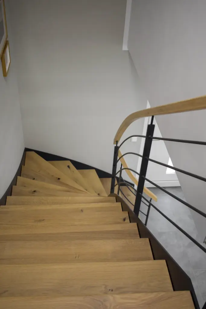 Eine Holztreppe mit einem Handlauf aus schwarzem Metall und Holz führt nach unten zu einem gefliesten Boden. Die Wand ist hell, und an der linken Seite hängen Bilderrahmen. Der Raum ist durch natürliches Licht gut beleuchtet.