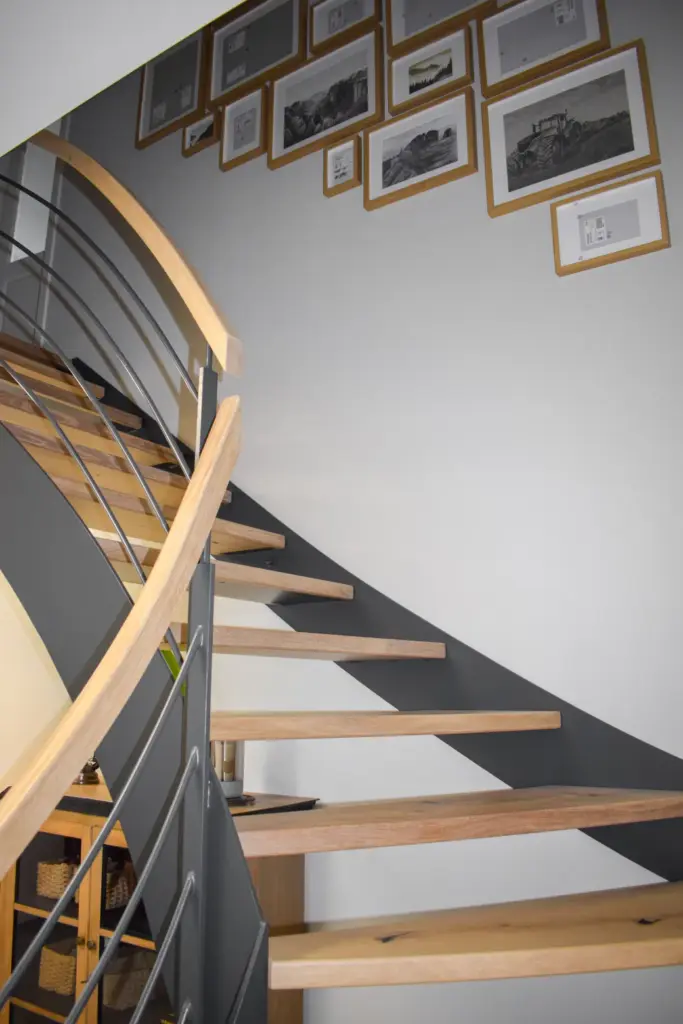 Eine moderne Holztreppe mit Metallgeländer schlängelt sich neben einer Wand nach oben, die mit gerahmten Schwarz-Weiß-Fotografien und Kunstwerken in einem Rastermuster dekoriert ist. Unter der Treppe sind Weidenkörbe zu sehen.