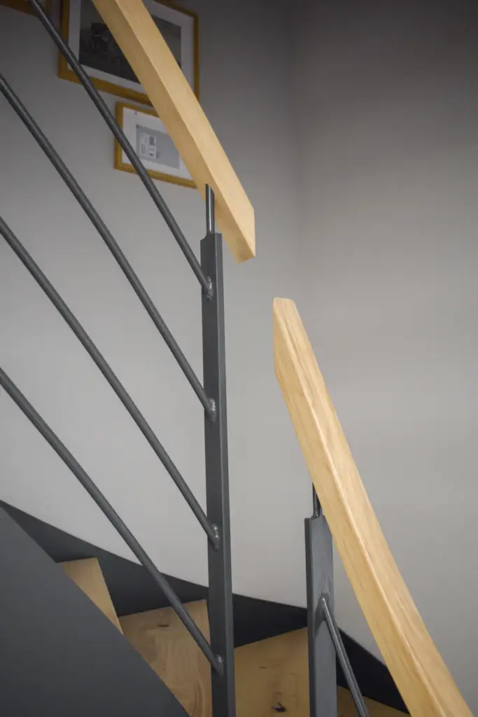 Eine Nahaufnahme einer modernen Treppe mit Handläufen aus hellem Holz und dunklen Metallgeländern. Gerahmte Bilder hängen an der grauen Wand im Hintergrund.