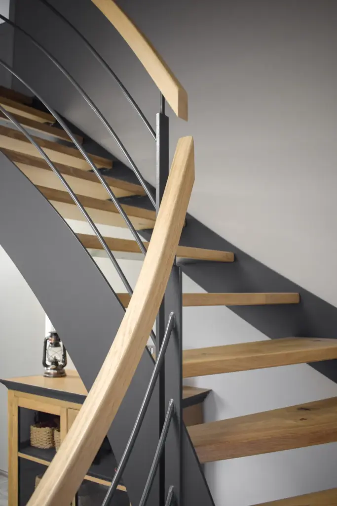 Moderne Treppe mit Holzstufen, glattem grauem Metallrahmen und einem hölzernen Handlauf vor einer hellgrauen Wand. Unter der Treppe befindet sich ein kleiner Schrank mit einer Laterne.