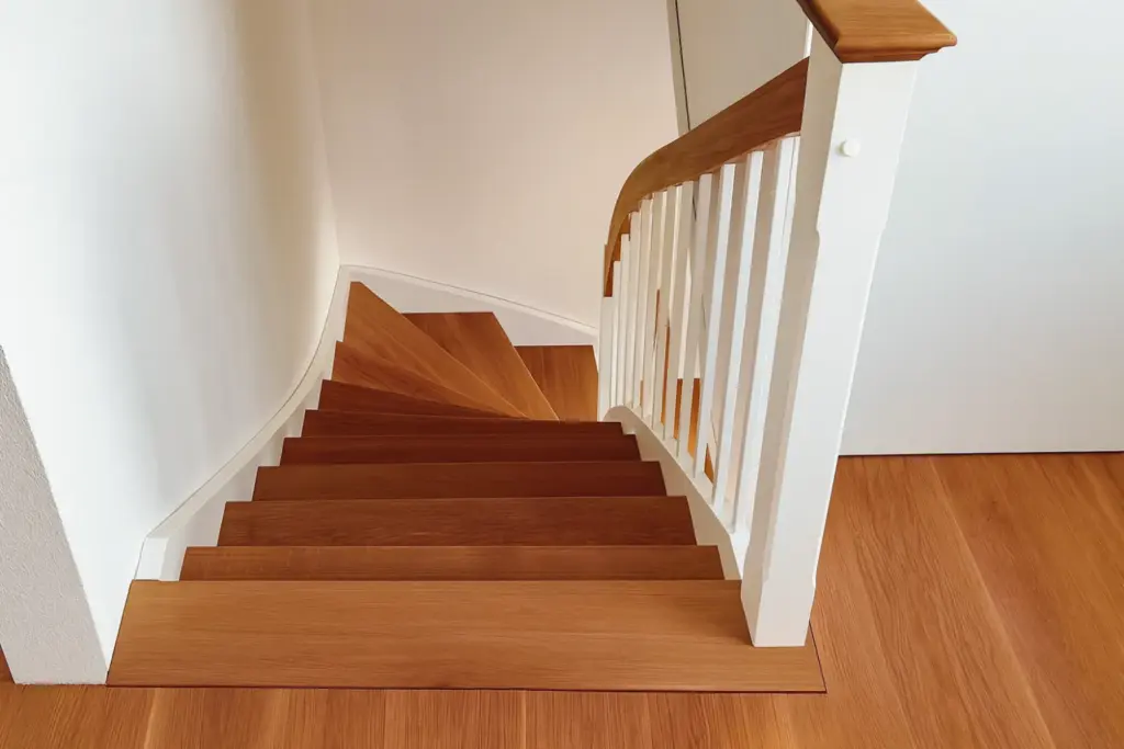 Eine Holztreppe mit weißem Geländer schlängelt sich nach unten, mit polierten braunen Stufen und passenden Parkettböden in einem hellen, sauberen Innenraum.