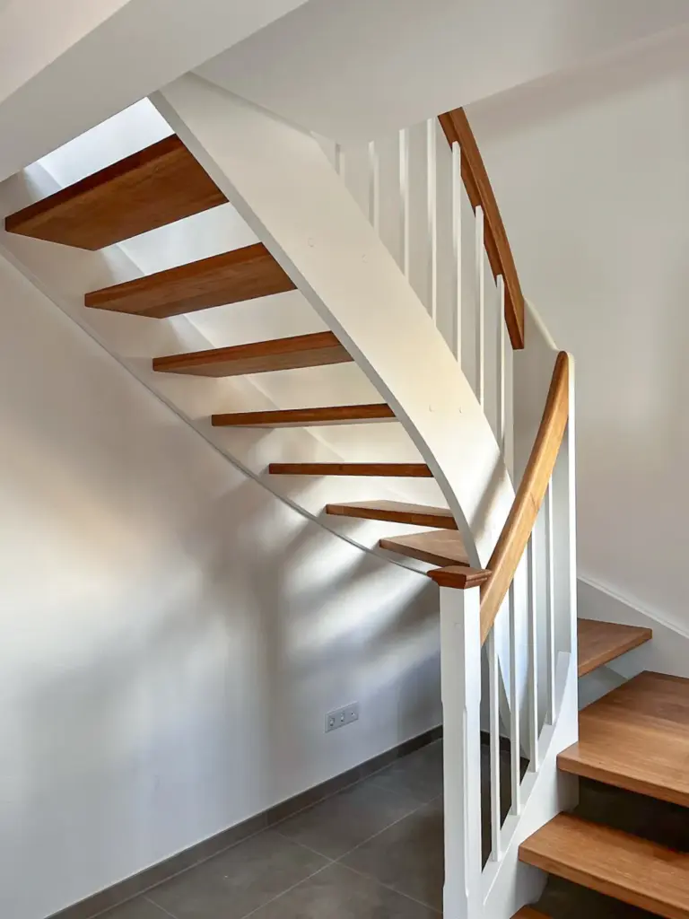 Eine moderne Holztreppe mit weißem Geländer und offenen Setzstufen wird in einem hellen Raum mit gefliestem Boden gezeigt. Das Sonnenlicht wirft Schatten auf die Wand unter der Treppe.