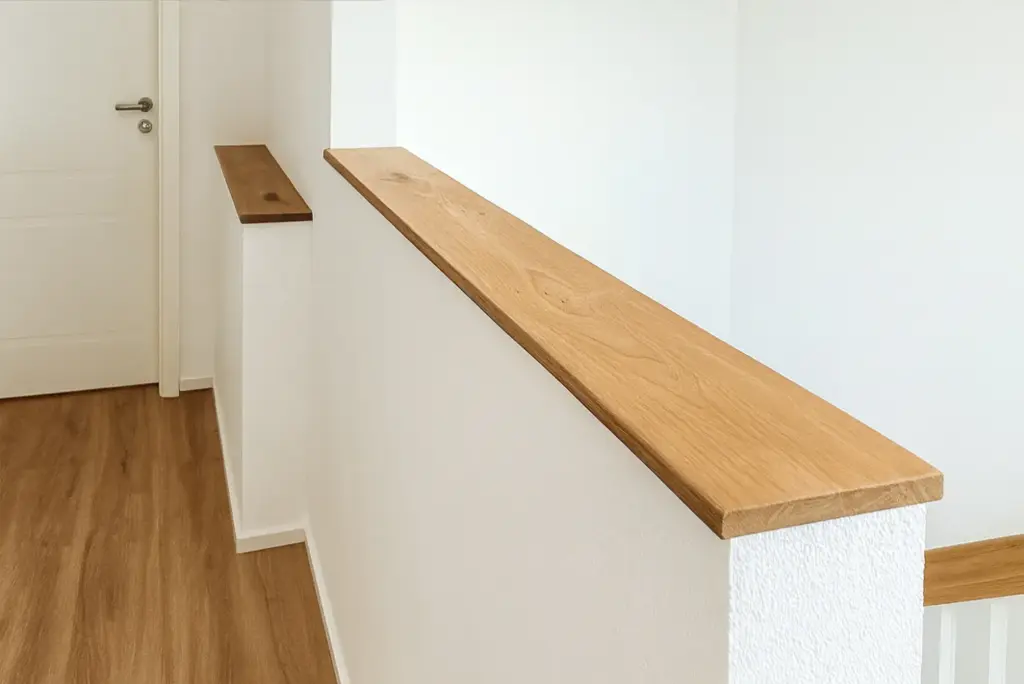 Ein moderner Flur mit weißen Wänden, hellem Holzfußboden und einer weißen Halbwand mit einem glatten Geländer aus Naturholz, das zu einer geschlossenen weißen Tür führt.
