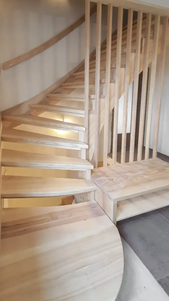 Daneben verläuft eine moderne Holztreppe mit offenen Setzstufen und vertikalen Lamellen, die sich nach links nach oben wölbt, mit natürlichen Holztönen und minimalistischem Design.