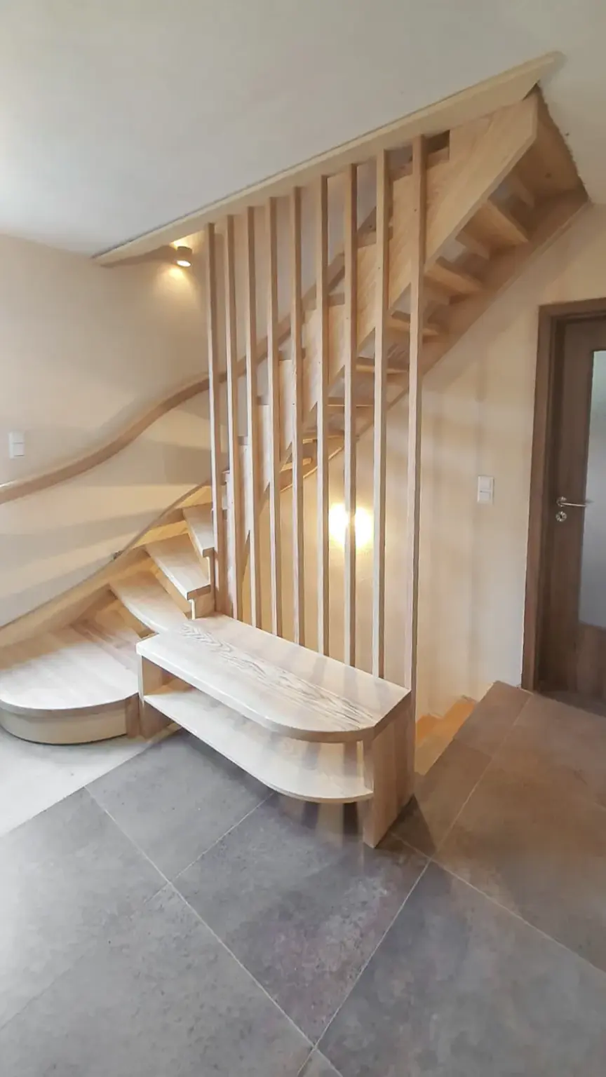 Eine moderne Holztreppe mit offenen Setzstufen und vertikalen Latten als Geländer, mit geschwungenen Stufen am unteren Ende und einer passenden Holzbank, vor beigen Wänden und grauem Fliesenboden.