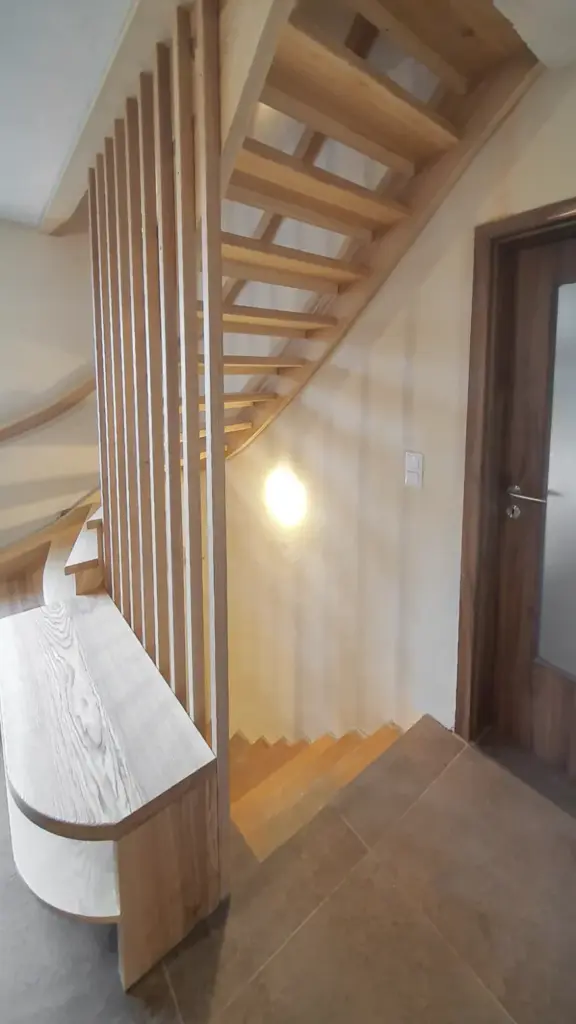 Moderne Holztreppe mit offenen Setzstufen und geschwungenem Design, daneben vertikale Holzlatten und eine eingebaute Bank. Der Bereich ist gut beleuchtet und verfügt über einen neutralen Fliesenboden und eine Holztür.