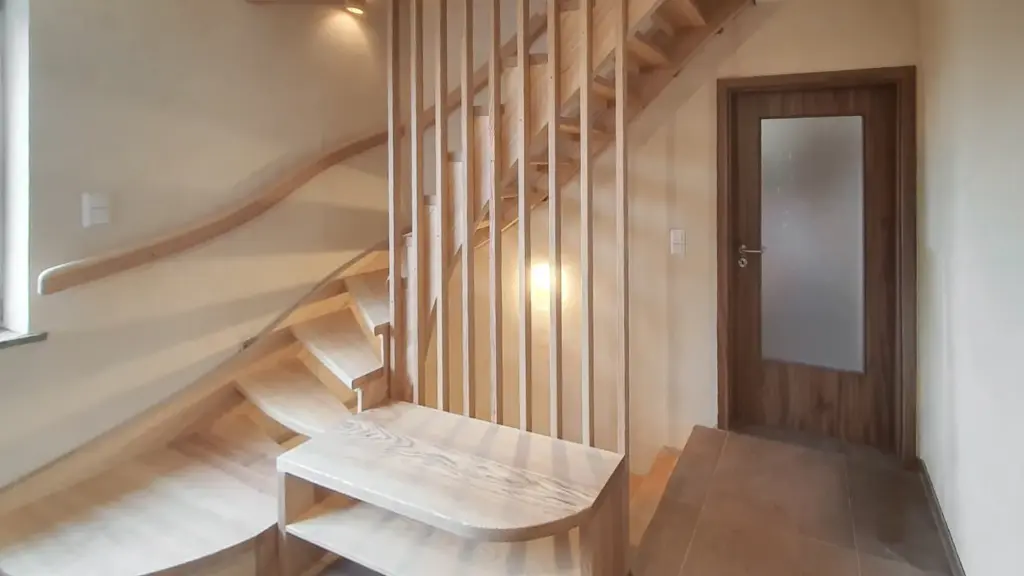 Holztreppe mit vertikalen Latten und geschwungenem Handlauf neben einer Milchglastür; am Fuß der Treppe steht eine kleine Holzbank, und durch ein Fenster auf der linken Seite fällt Licht ein.