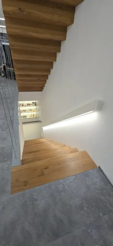 Eine moderne Holztreppe mit offenen Setzstufen führt an einer weißen Wand mit einer schlichten horizontalen Leuchte hinunter; am unteren Ende sind Regale mit verschiedenen Gegenständen zu sehen. Der Boden ist mit grauen Fliesen ausgelegt.