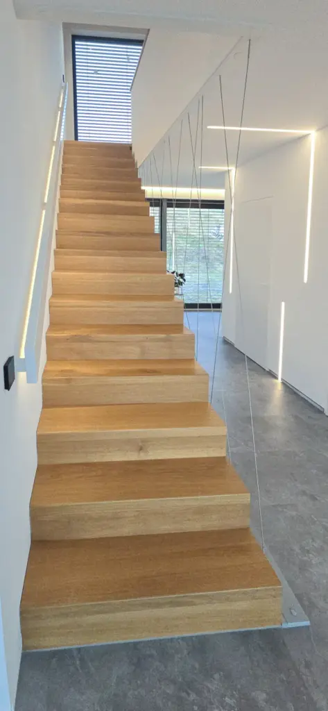 Eine minimalistisch gestaltete Holztreppe mit Kabelgeländer und Wandbeleuchtung führt nach oben in einen modernen, weiß getäfelten Innenraum mit grauem Fliesenboden. Große Fenster sorgen für natürliches Licht im oberen Bereich.