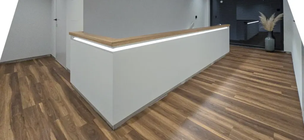 Ein moderner, minimalistischer Empfangstresen mit Holzplatte und weißer Front in einem Raum mit holzgemustertem Boden und neutralen Wänden; im Hintergrund steht eine hohe Vase mit Pampasgras.