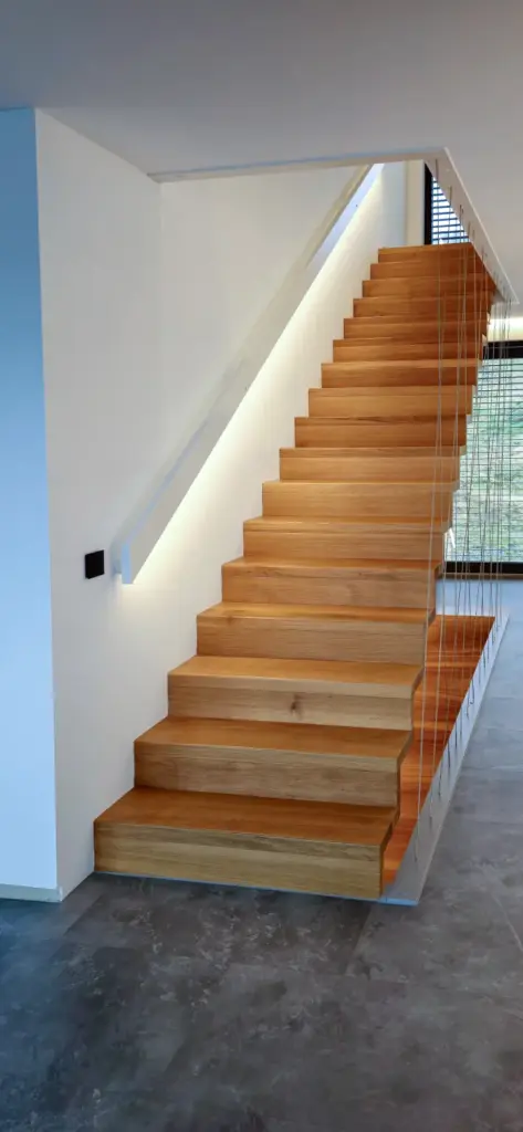 Moderne Holztreppe mit klaren Linien, offenen Setzstufen und Kabelgeländer; vor weißen Wänden mit einem LED-Beleuchtungsstreifen entlang des Geländers in einem modernen Wohnhaus mit grauem Fliesenboden.