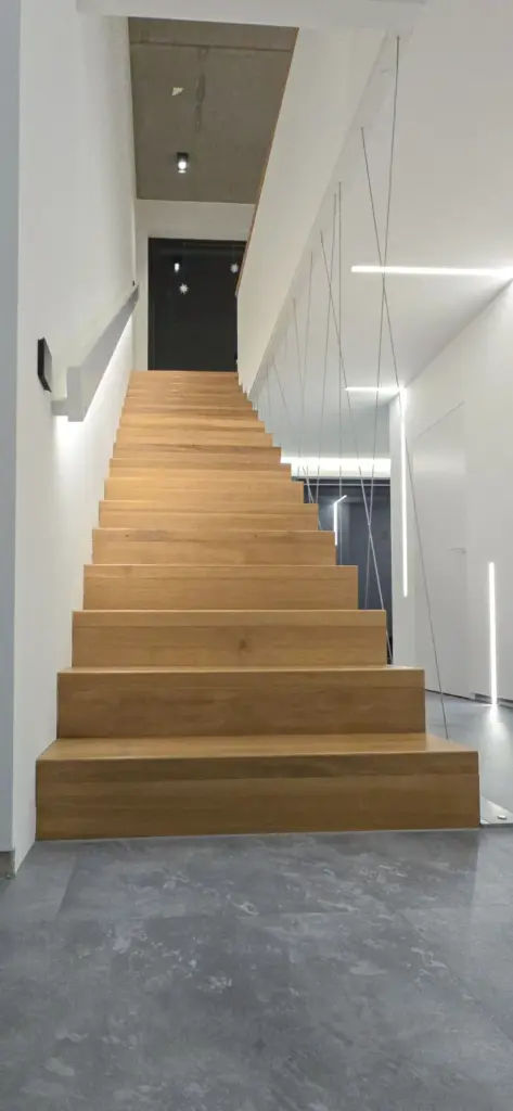 Eine moderne, minimalistische Treppe mit Holzstufen und dünnem Seilgeländer führt in einem hellen, weißen Flur mit grauem Bodenbelag und Deckeneinbauleuchten nach oben.