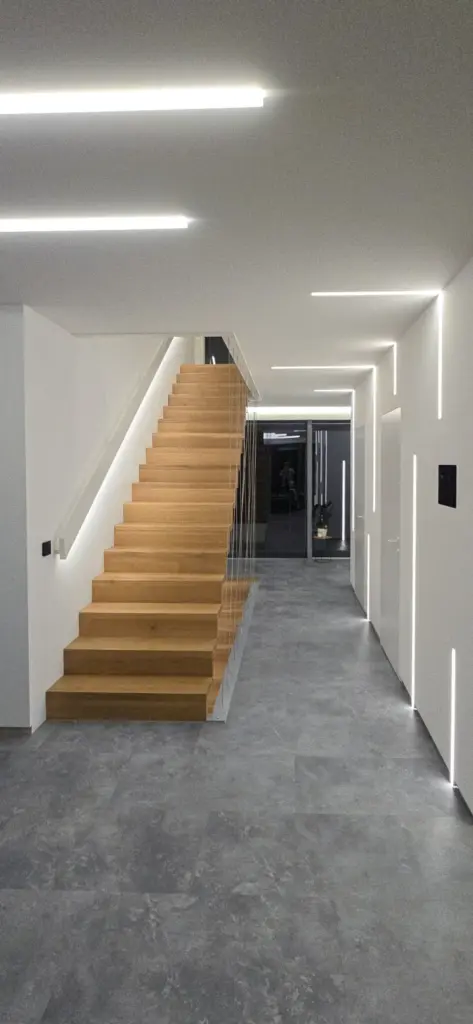 Modernes Interieur mit einer breiten Holztreppe mit Glasgeländer, grauem Steinboden und weißen Wänden, die durch schlichte, lineare LED-Leuchten an Decke und Wänden akzentuiert werden. Ein Flur führt zu einer Glastür am anderen Ende.