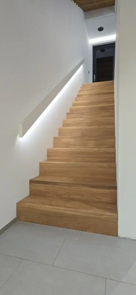 Eine moderne Innentreppe mit Holzstufen und weißem Handlauf an der linken Wand, die von einem LED-Lichtband beleuchtet wird und zu einer geschlossenen dunklen Tür am oberen Ende führt.
