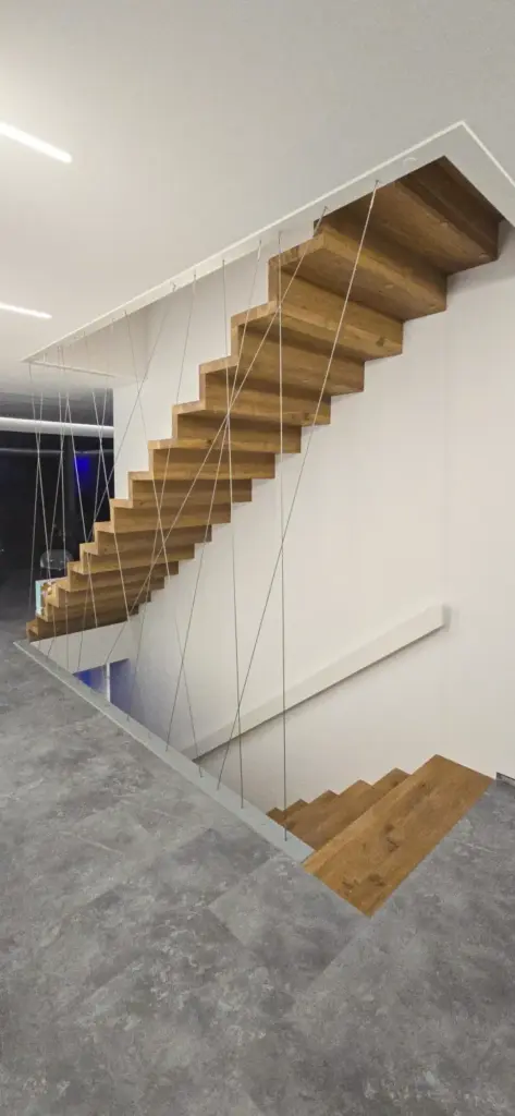 Eine moderne schwebende Holztreppe mit Metallseilen als Stütze führt in einem minimalistischen Interieur mit grauen Fliesenböden und weißen Wänden nach oben.