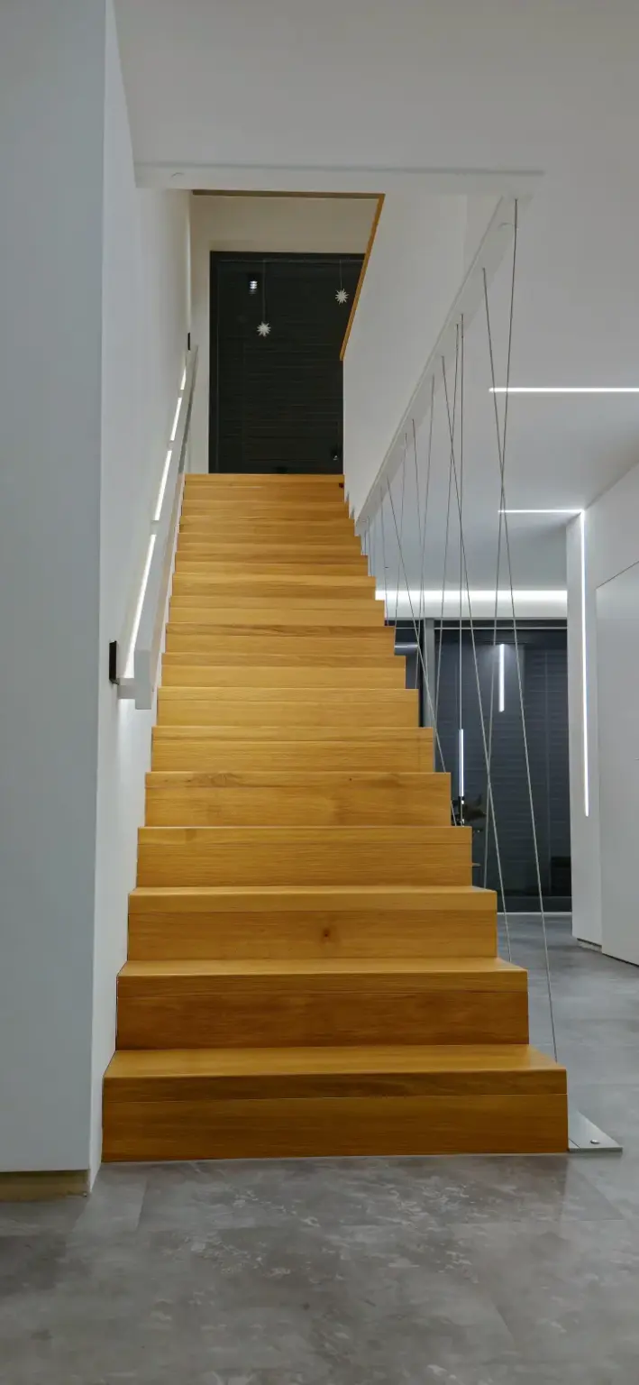 Eine moderne Holztreppe mit minimalistischem Metallgeländer führt zu einem Treppenabsatz in einem hellen, zeitgenössischen Interieur mit grauem Fliesenboden und schlichter, vertiefter Beleuchtung.