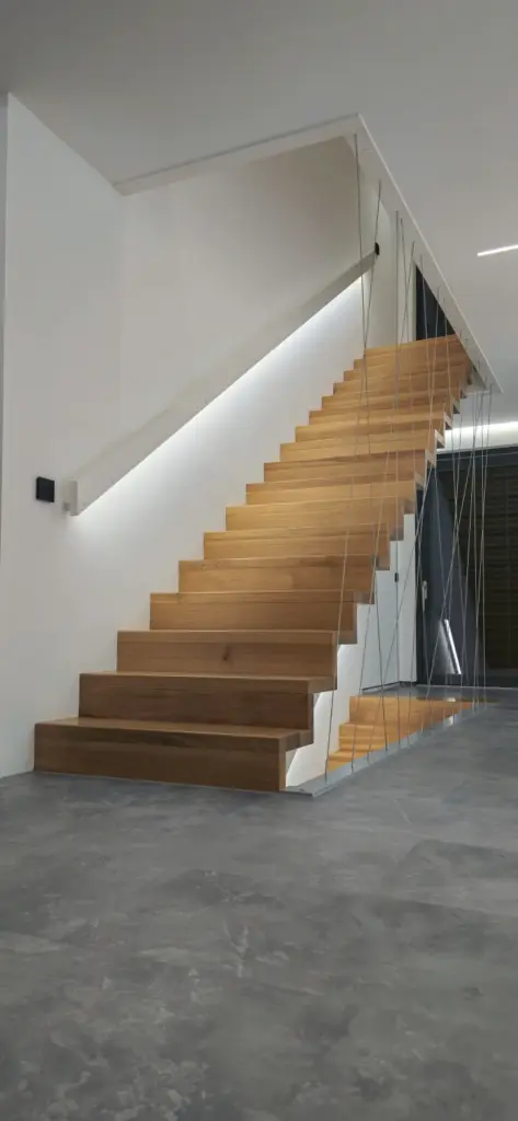 Eine moderne Holztreppe mit offenen Setzstufen, minimalistischem weißem Geländer und dünnen Metallseilen vor einer weißen Wand in einem schlichten, modernen Interieur mit grauem Betonboden.