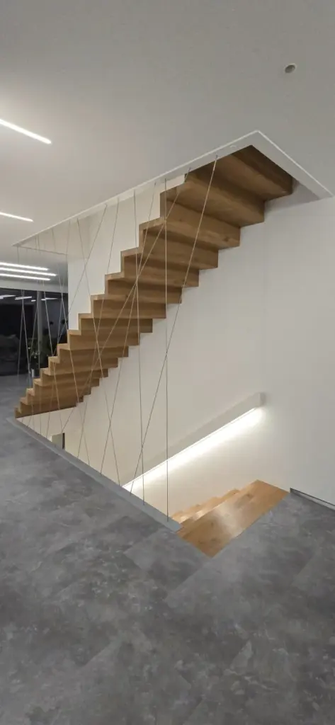 Moderne schwebende Holztreppe mit dünnen Metallseilen als Geländer, vor weißen Wänden und grauen Fliesenböden in einem minimalistischen, hellen Innenraum.