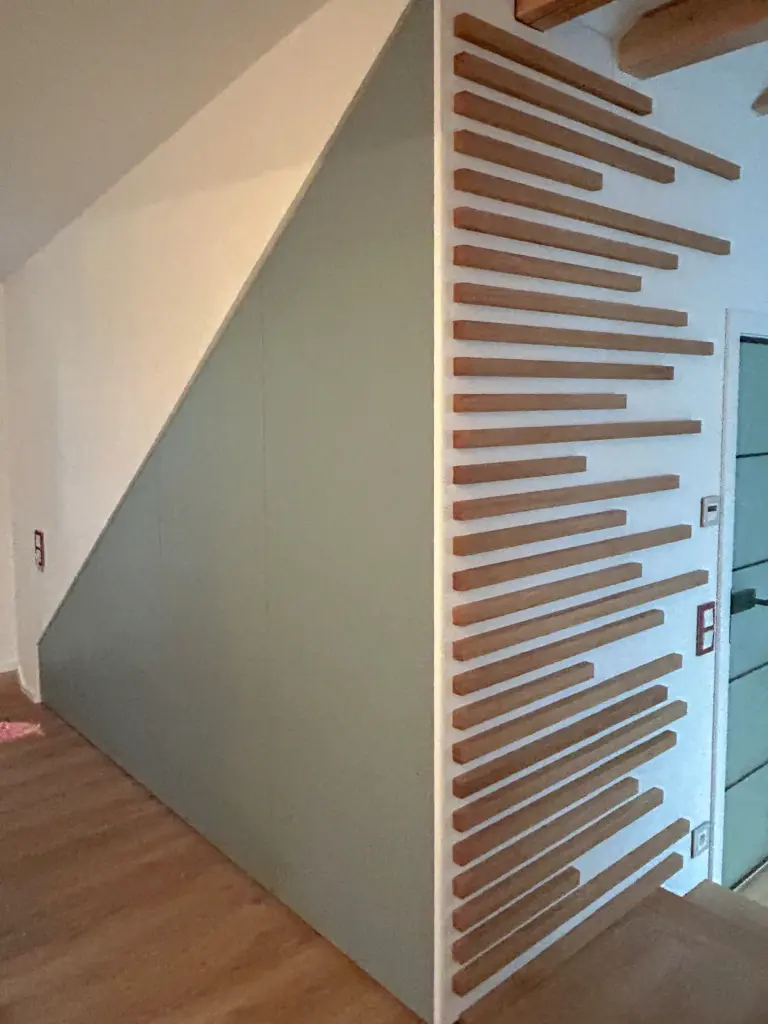 Eine moderne Treppe besteht aus einer schrägen Wand mit horizontalen Holzlatten unterschiedlicher Länge, die in einem versetzten Muster angeordnet sind, wodurch ein geometrisches Muster entsteht. Die Treppe und der Boden sind aus Holz gefertigt.