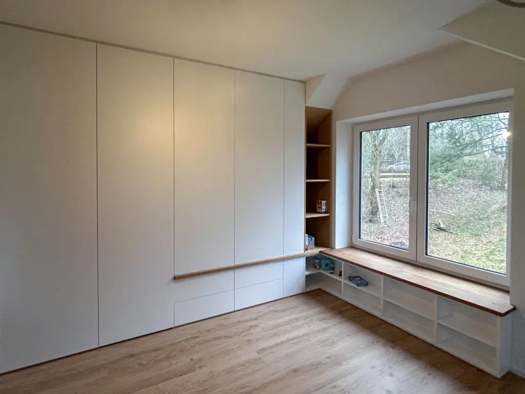 Ein minimalistischer Raum mit weißen Einbauschränken, einer Holzbank unter einem großen Fenster, offenen Regalen unter der Bank und Blick auf Bäume und eine Schaukel draußen. Der Boden ist aus hellem Holz.